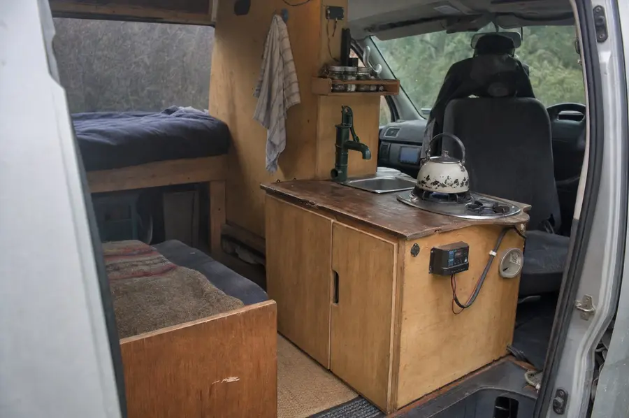 Fitted timber cabinetry, small kitchen bench and made-up bed inside a converted campervan parked in New Zealand
