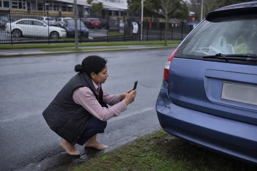 Woman photographing a dented rear bumper with her phone on a quiet Auckland street