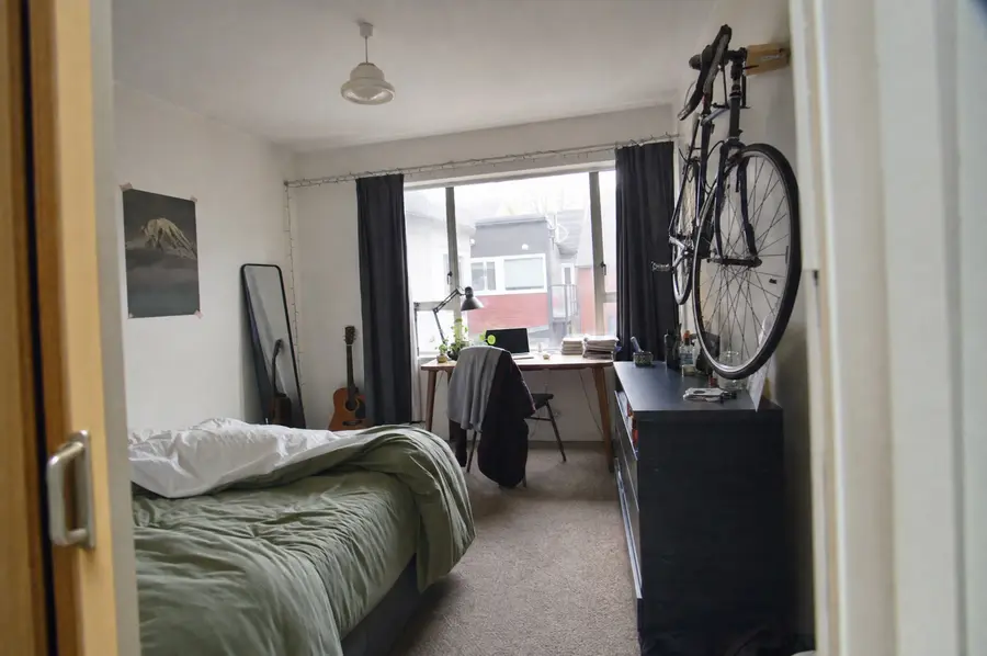 Bedroom in a rented Wellington flat with a made bed, clothes rack, guitar and laptop on the duvet
