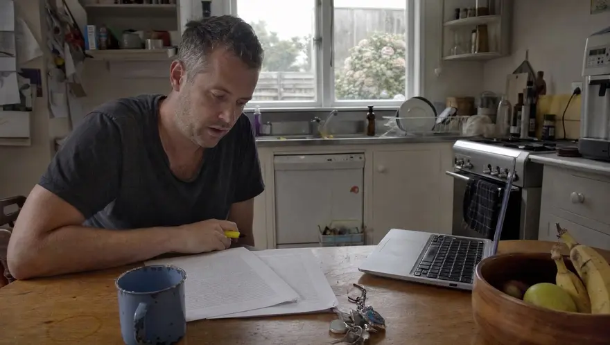 Man at a kitchen table comparing two printed car insurance policy documents side by side with a highlighter