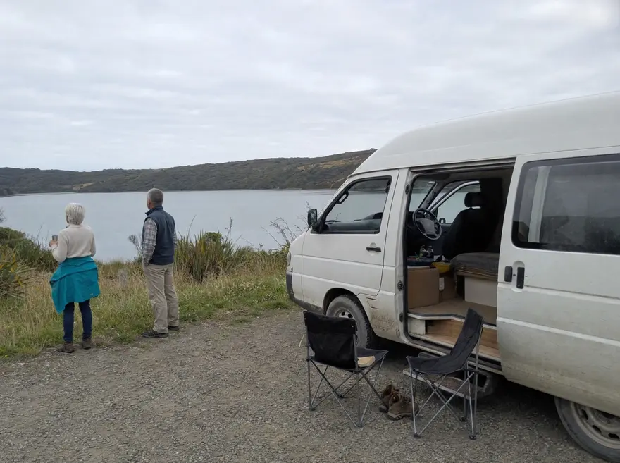 Couple in their sixties beside a white campervan parked at a gravel pull-in overlooking a New Zealand coastal inlet