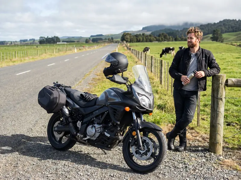 Motorcycle rider stopped on a quiet New Zealand rural highway with rolling green farmland behind