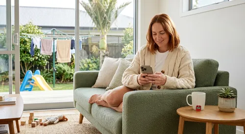 Woman on a sofa filling out an insurance quote on her phone in a sunny New Zealand living room