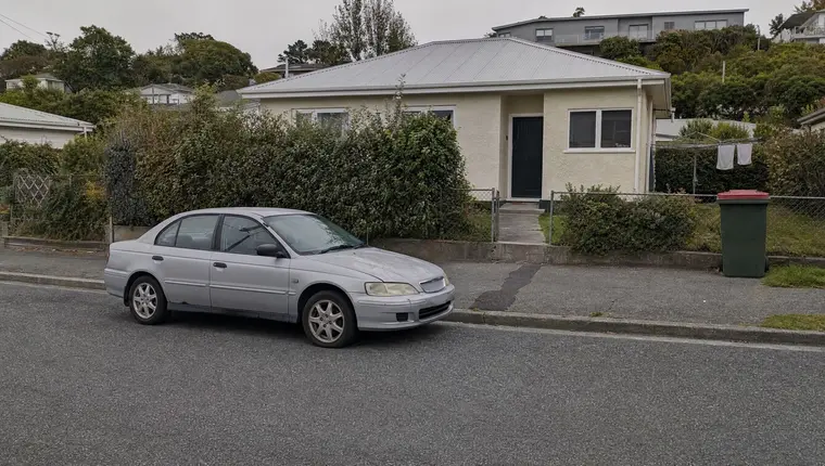 An older, well-used silver sedan parked at the kerb outside a modest Dunedin home