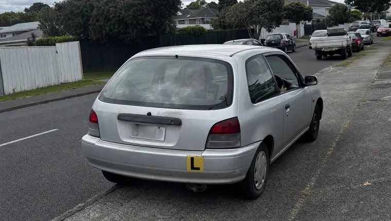 Learner driver at the wheel of a hatchback with a supervising adult in the passenger seat and a yellow L plate on the rear bumper